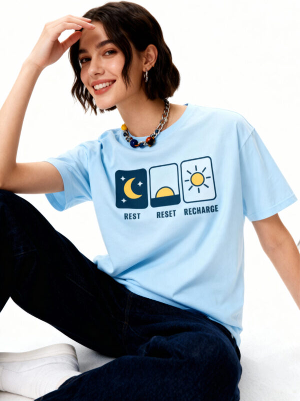 A brunette woman wearing a light blue 'Gildan 64000' t-shirt featuring a minimalist 'Rest, Reset, Recharge' graphic with moon and sun icons.