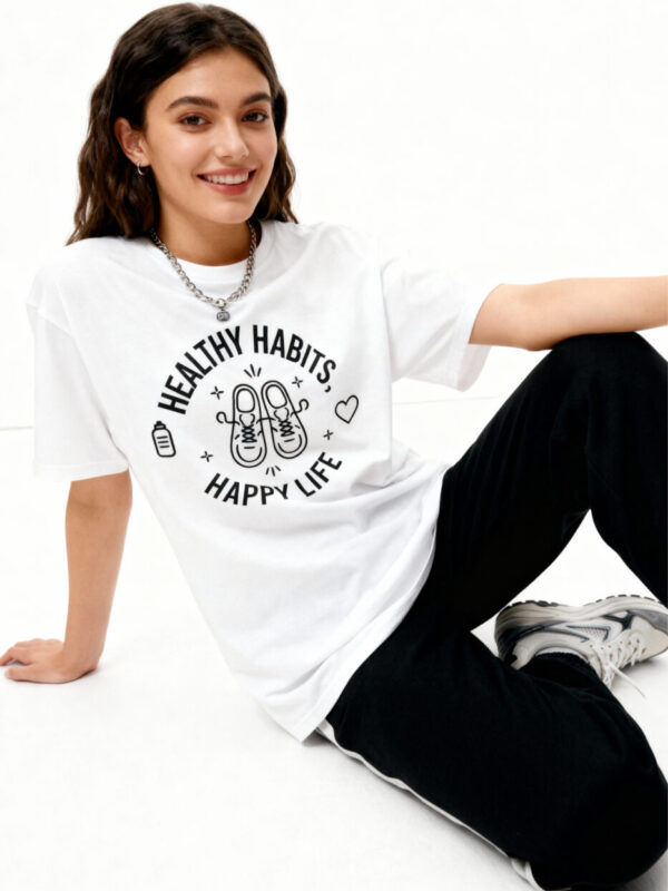 A brunette woman sitting and wearing a white 'Gildan 64000' t-shirt featuring a black 'Healthy Habits, Happy Life' graphic with trainers and a water bottle icon.