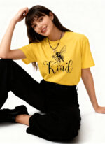Dark-haired model in yellow oversized "Kind" bee T-shirt and black pants, seated pose.