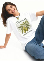 Smiling woman in a white Valencia graphic T-shirt and blue jeans sitting on the floor. The shirt features an illustration of olives on a branch with text.