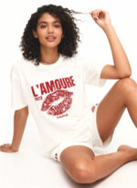 Woman sitting in a white studio wearing a white T‑shirt and white shorts with a red “L’AMOUR 1972 PARIS” lips graphic, holding a curl of her hair.