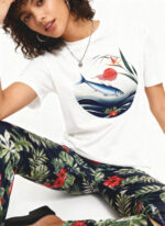 Woman sitting in a white studio wearing a white T‑shirt with a circular illustration of a fish swimming over waves beneath a red sun and flowers, paired with dark tropical floral-print pants and black shoes.