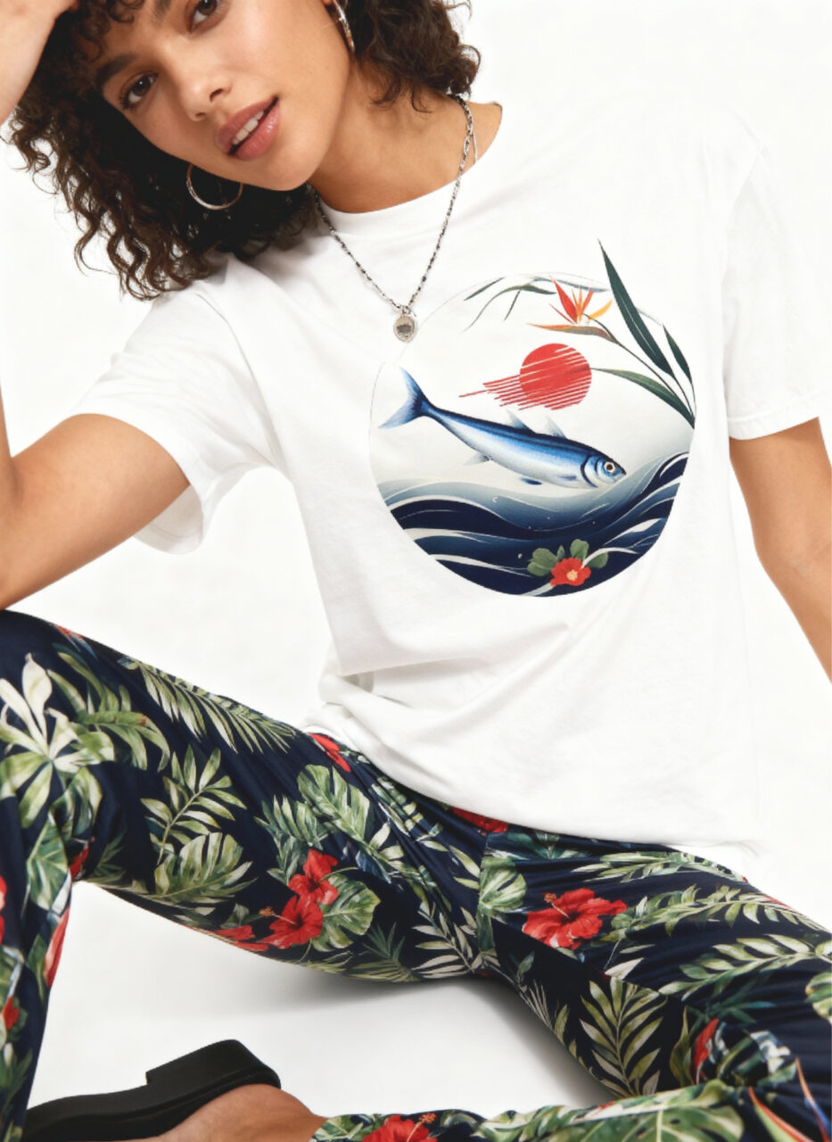 Japanese Sardine Art Woman sitting in a white studio wearing a white T‑shirt with a circular illustration of a fish swimming over waves beneath a red sun and flowers, paired with dark tropical floral-print pants and black shoes.