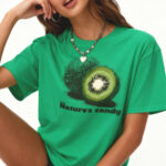 Person with long brown hair wearing a bright green T‑shirt featuring a kiwi graphic and the words “Nature’s candy,” posing with one hand resting against their face on a white background.