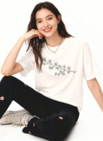 A full-length studio shot of a cheerful young woman with long, dark brown hair, sitting on a white floor. She is wearing a white graphic t-shirt featuring a black line drawing of a cherry blossom branch. She pairs it with black jeans embroidered with small pink flowers and white low-top sneakers. She is smiling warmly at the camera with her chin resting in her hand.